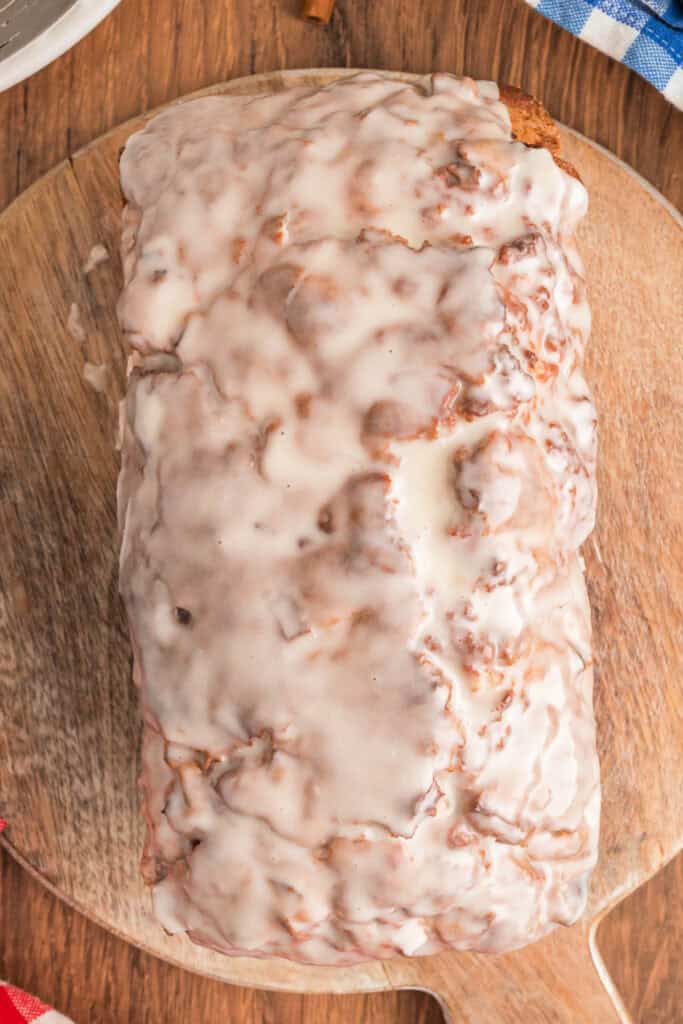 Apple fritter bread loaf covered in vanilla glaze, set on a wooden board with glaze dripping down the sides.