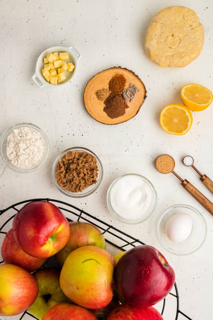 Overhead view of apple pie ingredients including apples, flour, sugars, lemon, butter, egg, and spices.