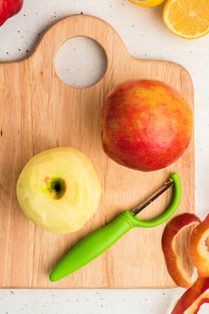 Fresh apple being peeled with a green peeler on a wooden cutting board.