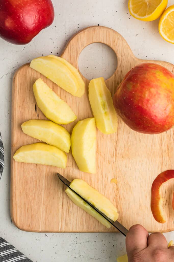 Apple wedges being cut into thinner slices with a knife on a cutting board.