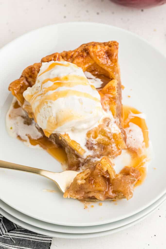 Slice of apple pie with melting ice cream and caramel drizzle on a plate.