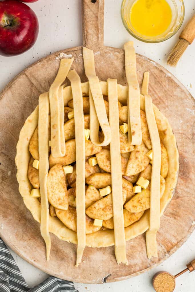 Apple pie with lattice strips being woven, egg wash and sugar nearby.