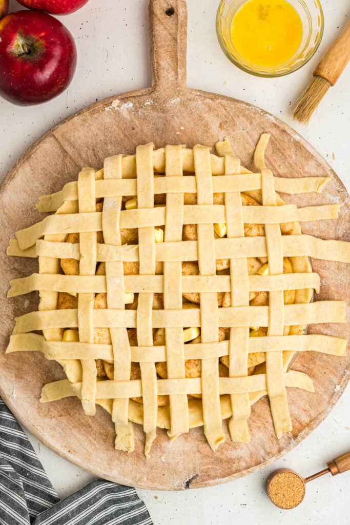 Unbaked lattice top apple pie before trimming edges, with egg wash and sugar nearby.