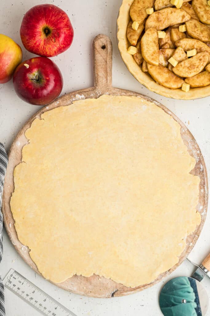 Rolled out pie dough on a wooden board next to filled apple pie crust.