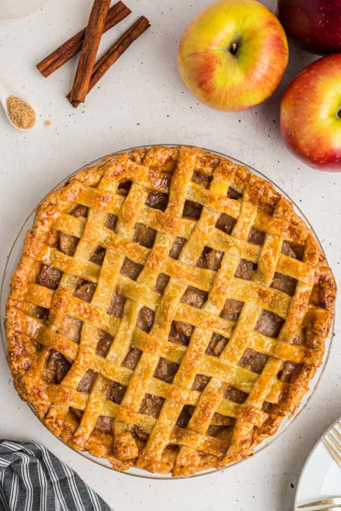 Golden baked apple pie with lattice crust on table with apples and cinnamon sticks.