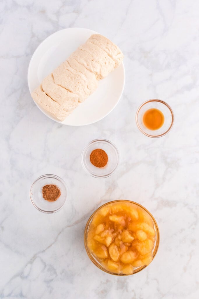 Ingredients for apple pie bombs including biscuit dough, apple pie filling, vanilla, and spices on a marble counter.