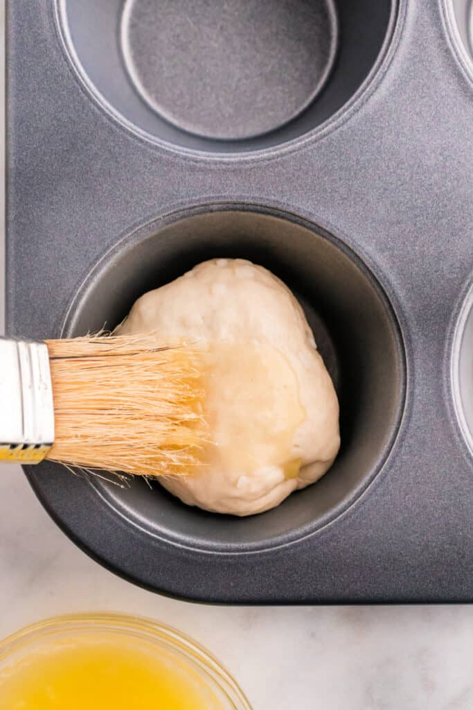 Brushing melted butter onto the top of an apple pie bomb before coating with cinnamon sugar.
