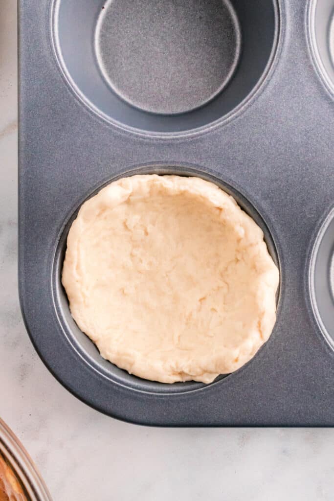 Biscuit dough pressed into a muffin pan cup to form a base for apple pie bombs.