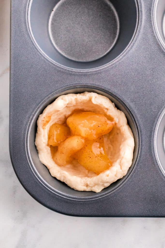 Biscuit dough cup filled with spiced apple pie filling in a muffin tin cavity.
