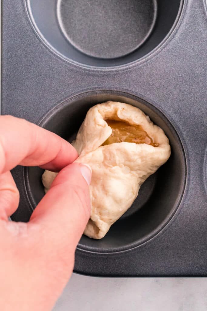 Hands folding biscuit dough over apple pie filling to form a sealed apple pie bomb.