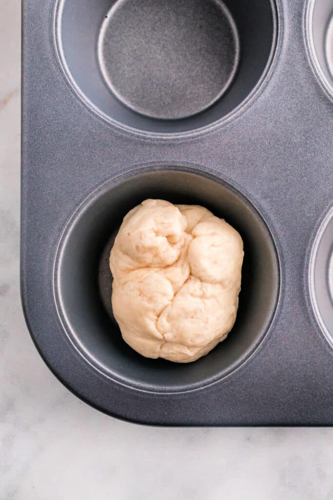 Sealed apple pie bomb dough ball sitting in a muffin pan cavity before baking.