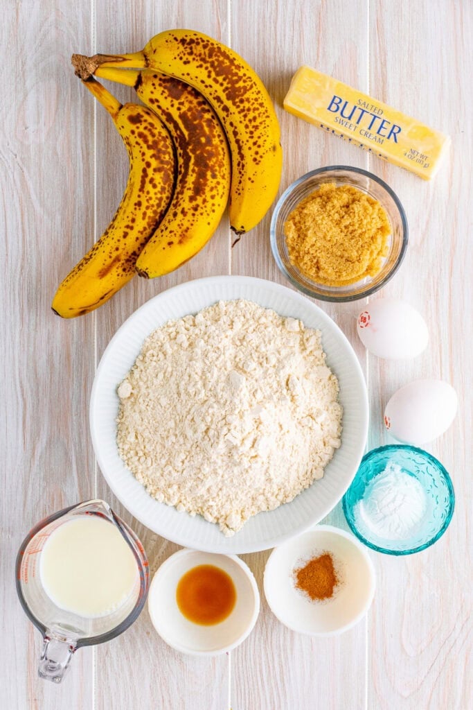 Overripe bananas, butter, brown sugar, flour, eggs, milk, and cinnamon displayed on a light wooden surface for banana fritters recipe