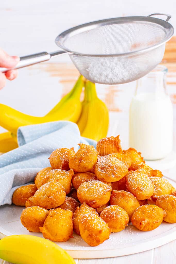 A pile of banana fritters being dusted with powdered sugar using a mesh strainer