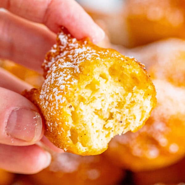 Close-up of a hand holding a soft, airy banana fritter dusted with powdered sugar