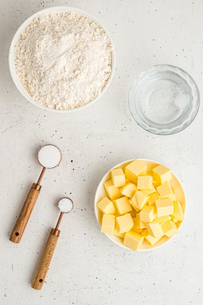 Overhead view of ingredients for butter pie crust including flour, butter, water, sugar, and salt