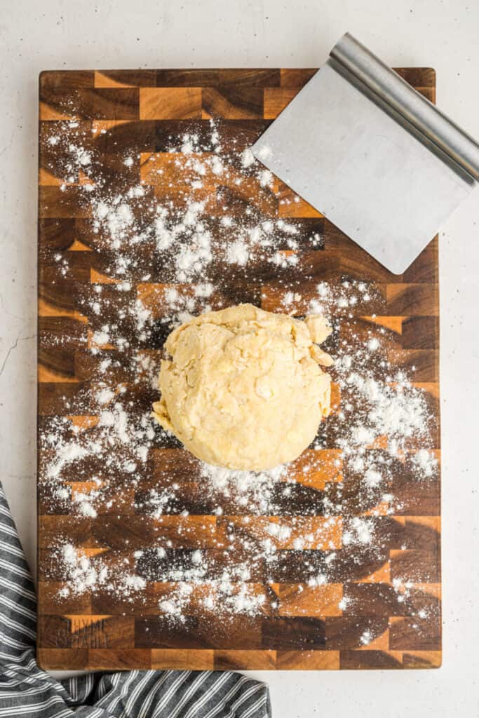 Ball of pie dough on a floured wooden board with a bench scraper beside it