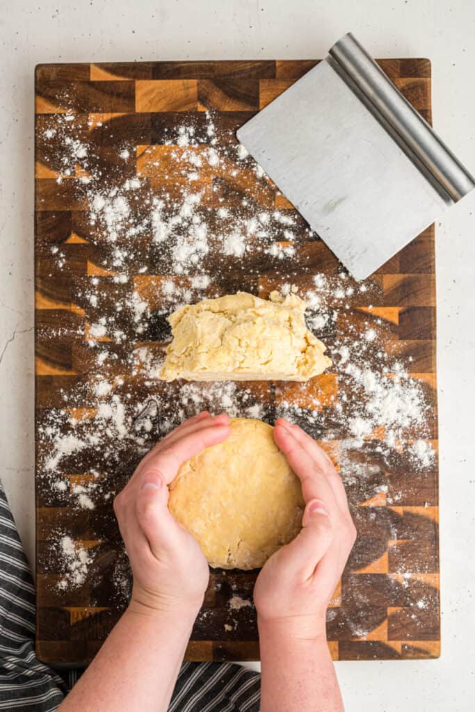 Hands shaping pie dough into a disc on a floured wooden board