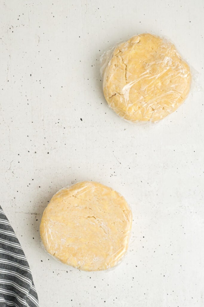 Two discs of butter pie crust dough wrapped in plastic wrap on a white surface