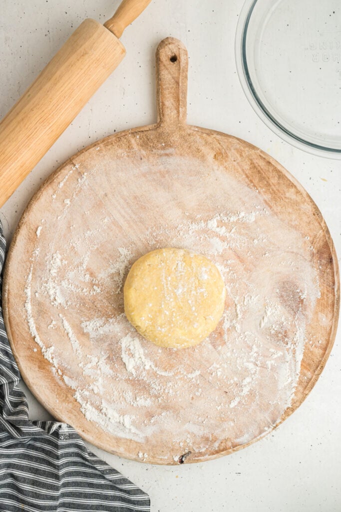 Disc of chilled dough on a floured wooden rolling board next to a rolling pin