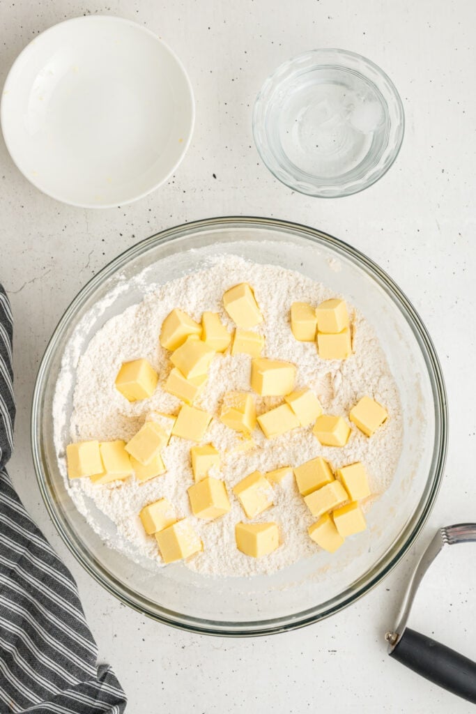 Cubed butter added to the flour mixture in a bowl with water and tools nearby
