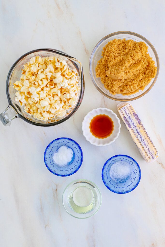 Overhead view of measured caramel corn ingredients including popcorn, brown sugar, vanilla, butter, salt, baking soda, and corn syrup in separate bowls