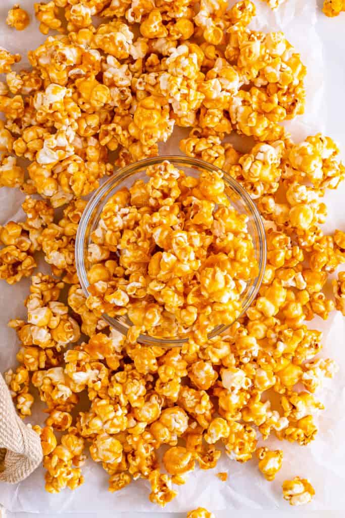 Overhead view of a clear bowl of caramel corn surrounded by loose popcorn on parchment paper
