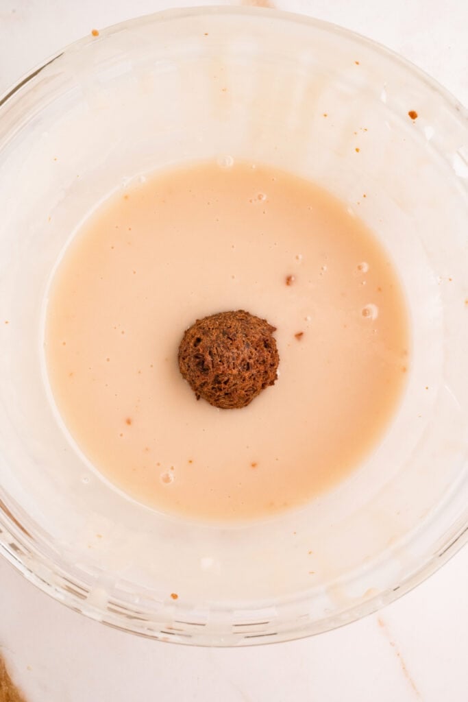 Fried donut hole dipped into vanilla glaze in a glass bowl