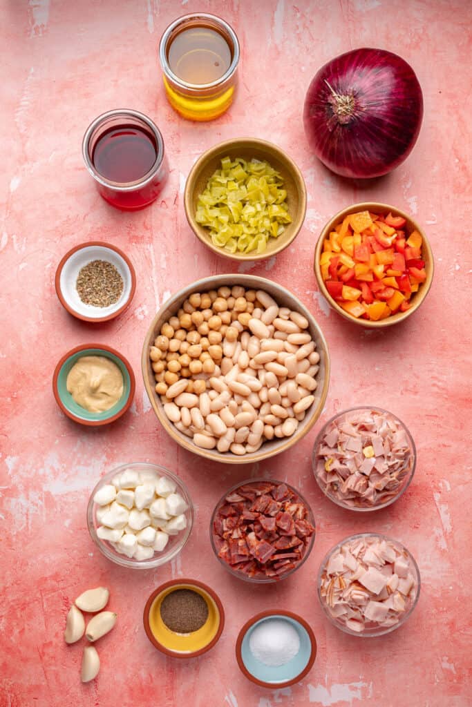 Overhead shot of salad ingredients including beans, diced peppers, pepperoncini, mozzarella, meats, and vinaigrette items on a pink background.