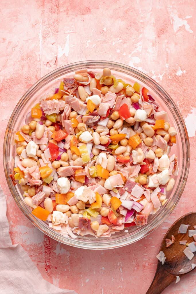 Grinder dense bean salad partially mixed in a glass bowl showing beans, meats, cheese, and veggies.