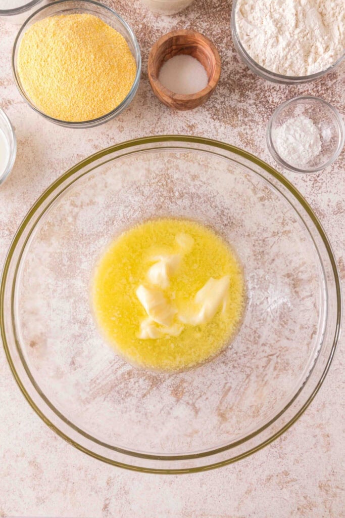 Mixing bowl with melted butter beginning to soften, surrounded by dry ingredients for cornbread.
