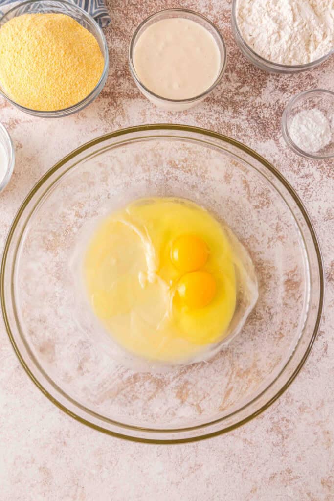Two eggs cracked into a bowl with melted butter and sugar mixture, surrounded by bowls of ingredients.