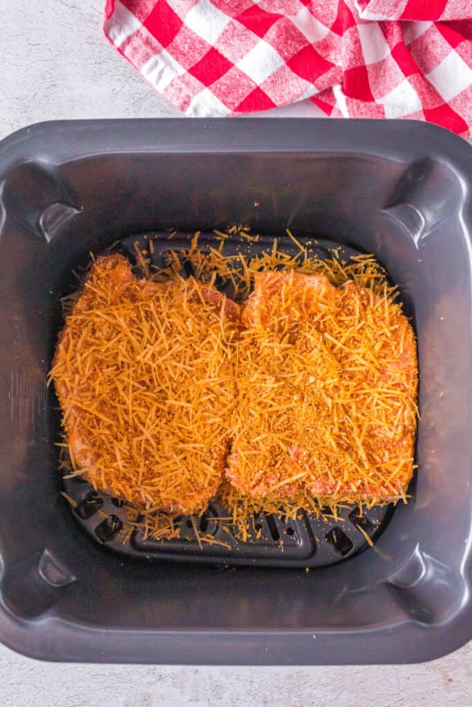 Two seasoned bone-in pork chops coated with Parmesan and crouton mixture placed in an air fryer basket ready to cook.