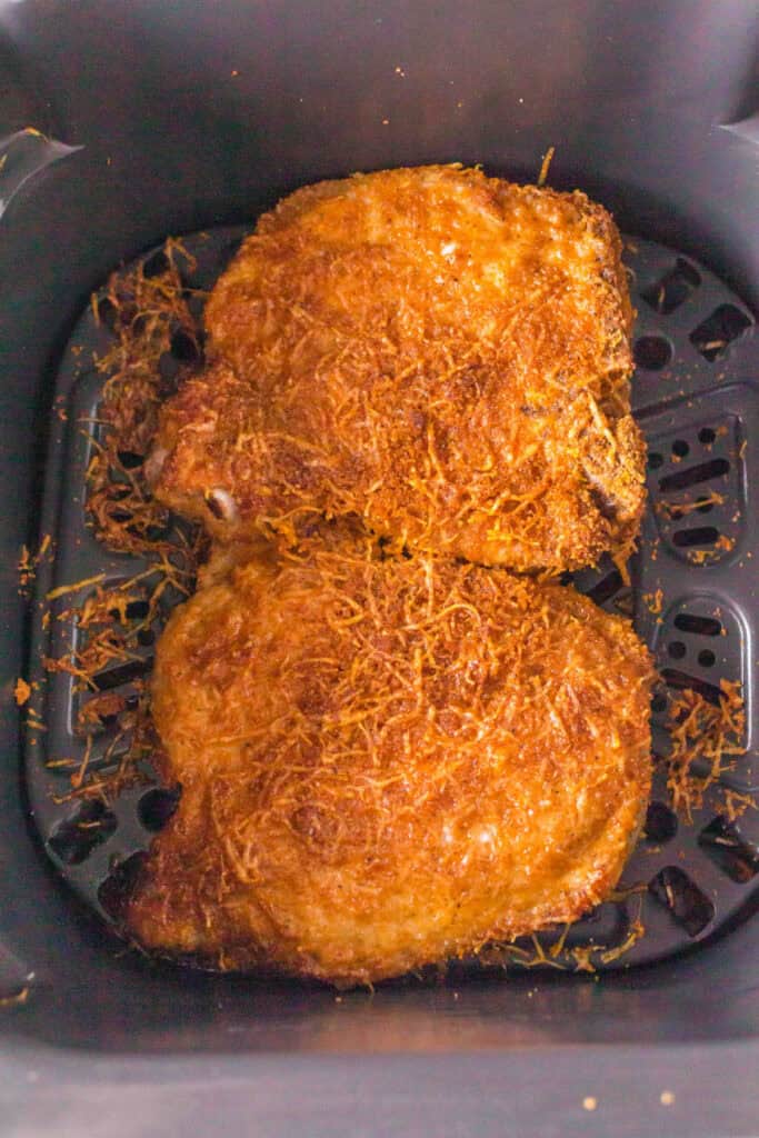 Two golden brown air fryer bone-in pork chops fresh out of the air fryer basket with crispy Parmesan crust.