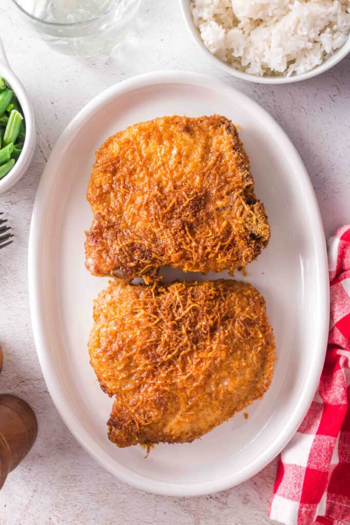 Two crispy bone-in pork chops served on a white platter with sides of rice and green beans in the background.