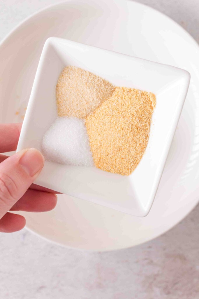 Small white dish with garlic powder, onion powder, and salt held over a mixing bowl.