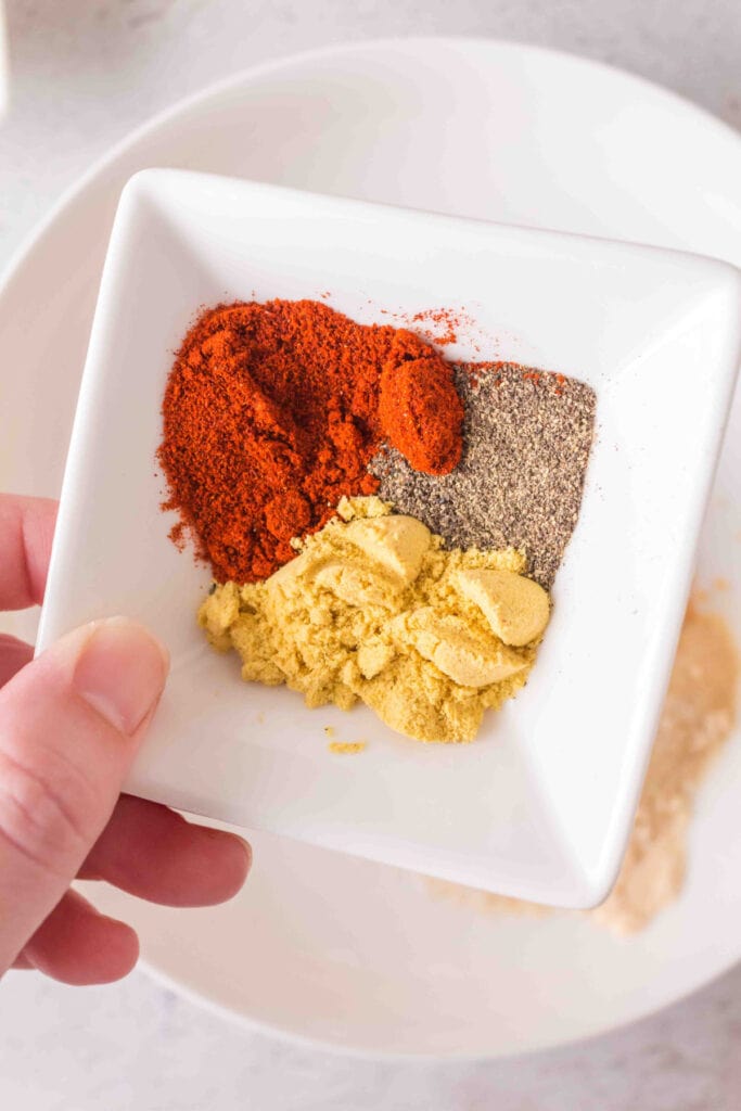 Small white dish with smoked paprika, ground mustard, and black pepper held above a bowl.