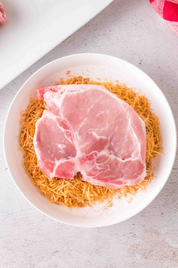 Raw pork chop placed in a bowl of seasoned Parmesan and crouton mixture before coating.