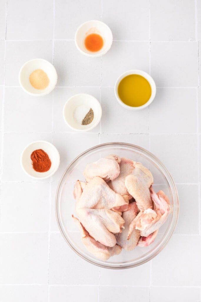 Raw chicken wings in a glass bowl with small bowls of olive oil, hot sauce, garlic powder, paprika, salt, and pepper on a white tile surface.