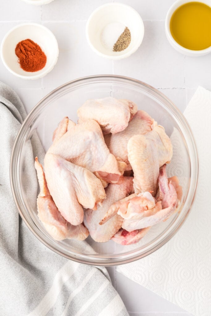 Close-up of raw chicken wings in a glass bowl with seasoning bowls nearby on a white tile counter with a striped kitchen towel.
