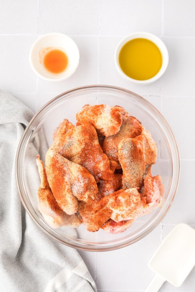 Raw chicken wings coated with garlic powder, paprika, salt, and pepper in a glass bowl with oil and hot sauce bowls beside it.