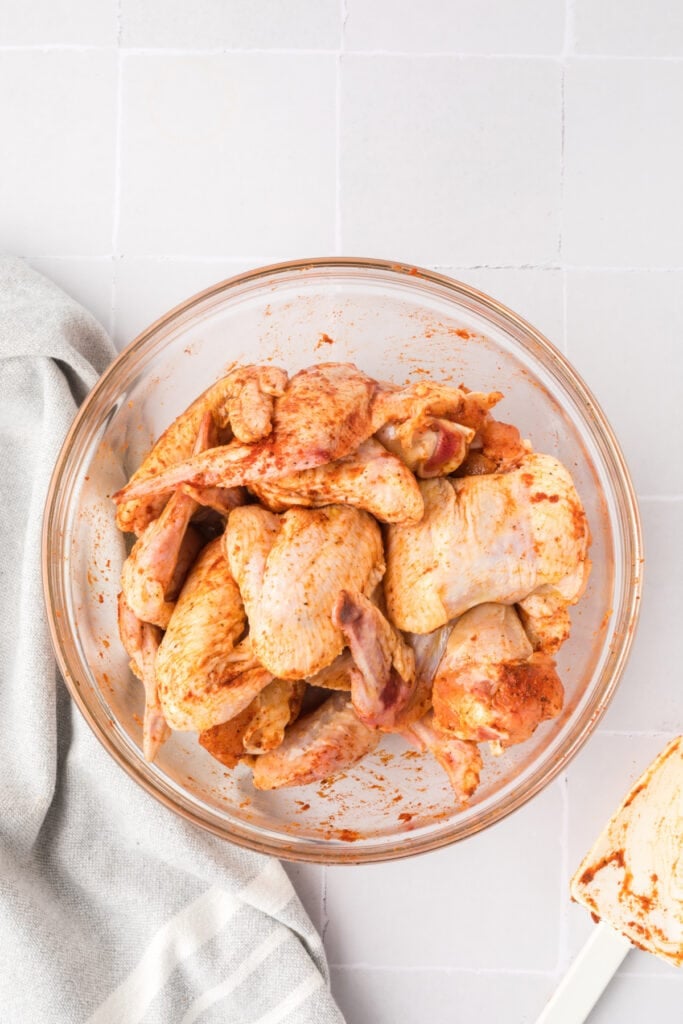 Chicken wings tossed with seasoning and hot sauce in a glass bowl with a spatula resting on the side.