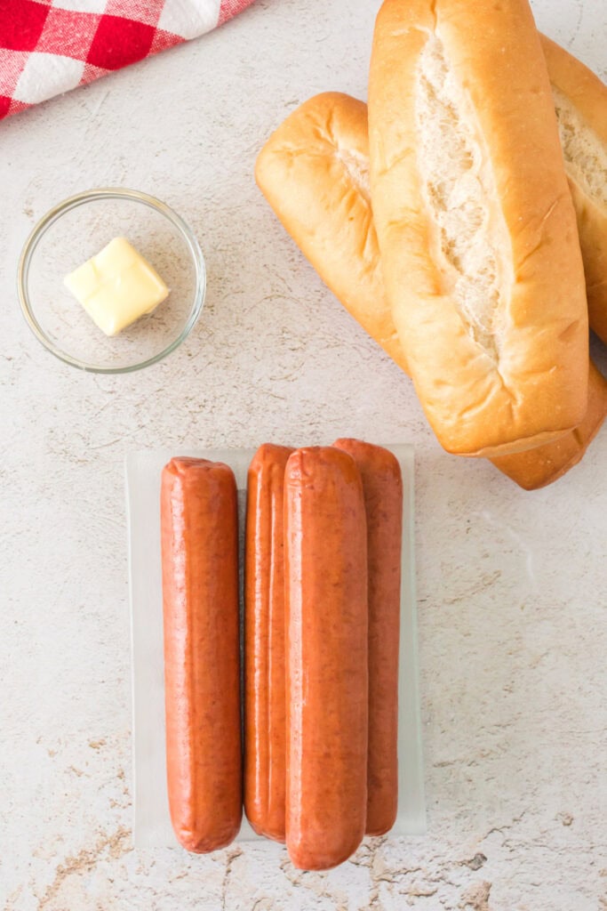 Raw hot dog sausages, hoagie buns, and a pat of butter on a light countertop