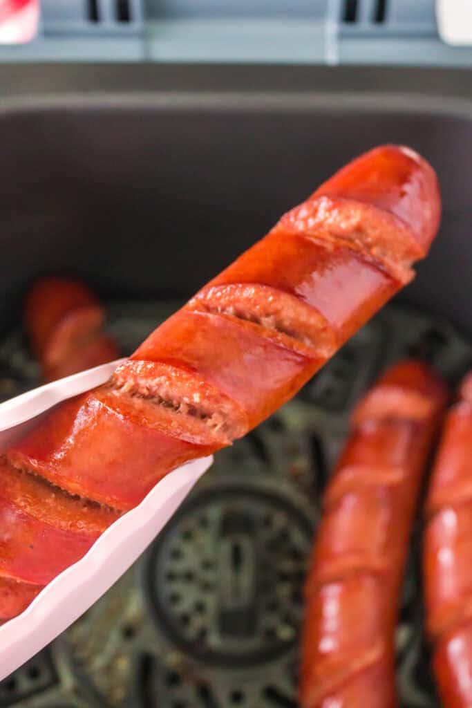 Tongs lifting a fully cooked scored hot dog from the air fryer