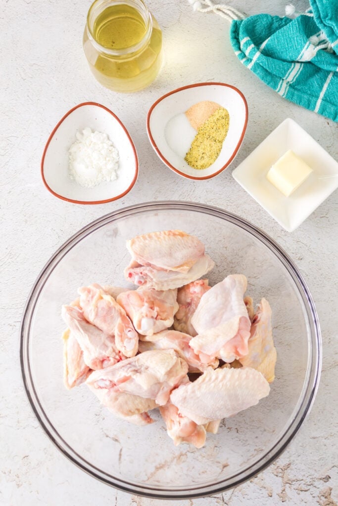 Overhead shot of raw chicken wings in a glass bowl with small bowls of cornstarch, seasoning, melted butter, and olive oil on the side.