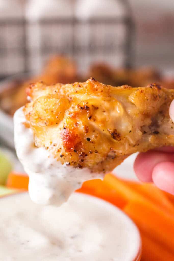Hand dipping a crispy lemon pepper chicken wing into ranch dressing with celery and carrots in the background.