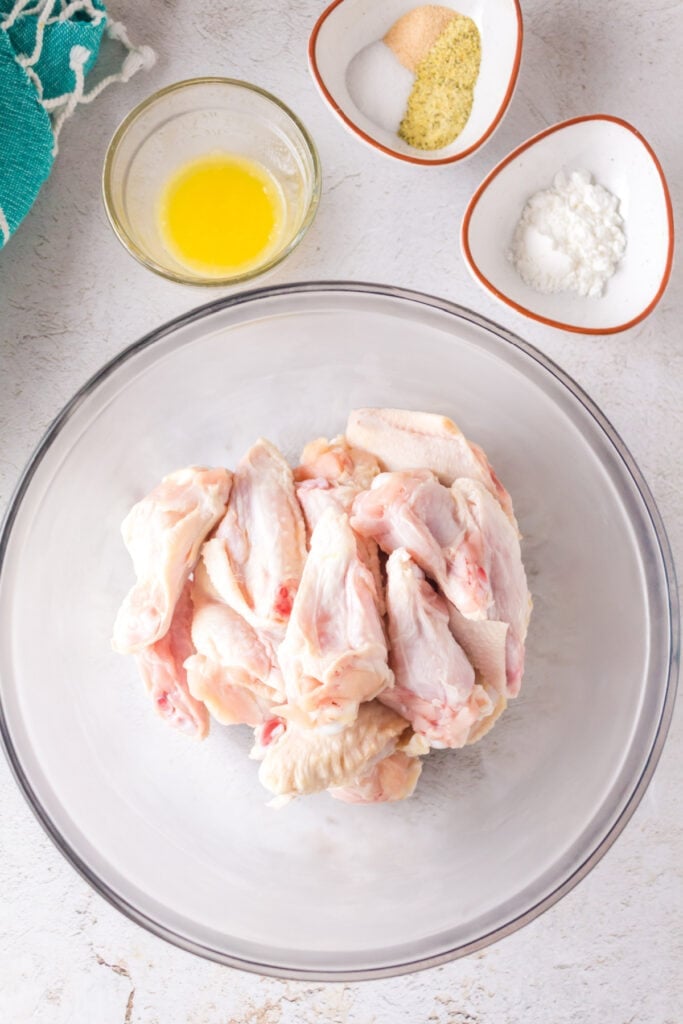 Raw chicken wings in a glass bowl with melted butter, seasoning mix, and cornstarch in small bowls nearby.