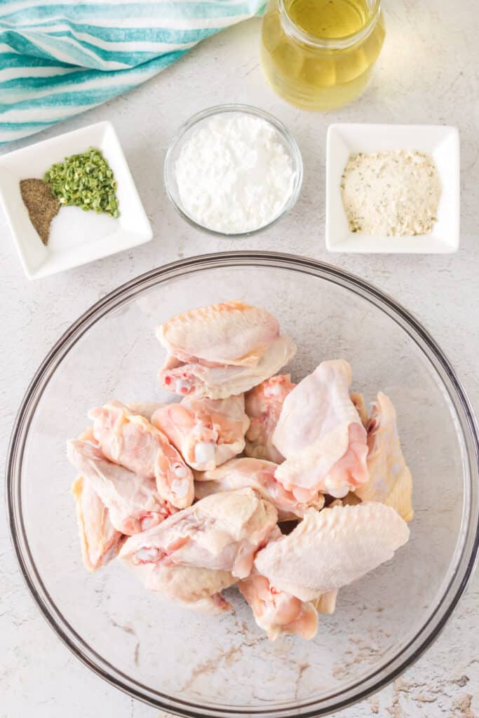 Raw chicken wings in a glass bowl surrounded by olive oil, cornstarch, seasoning mix, and spices on a white counter