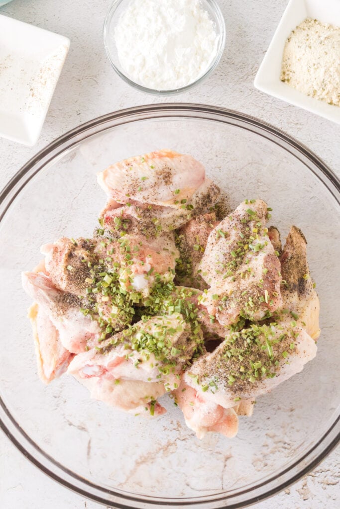 Chicken wings in a glass bowl sprinkled with salt, pepper, and chives before mixing