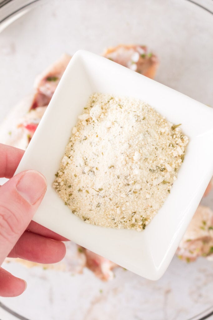 Hand holding a dish filled with ranch seasoning mix over raw chicken wings