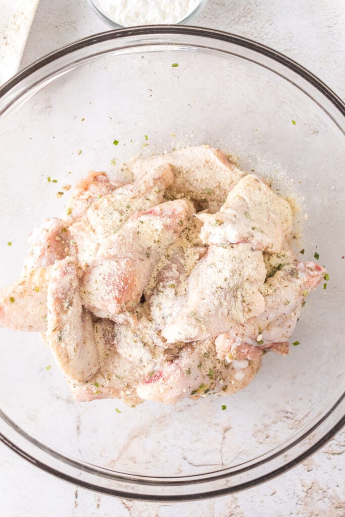 Raw chicken wings coated with ranch seasoning mix and herbs in a glass bowl